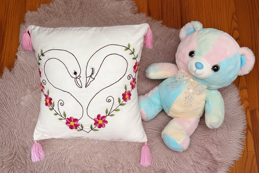 Decorative pillow with embroidered swans and flowers next to a colorful teddy bear on a textured surface.